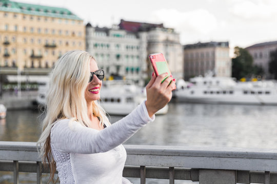 Portrait of the smiling young woman who is standing on the bridge and doing selfie photo