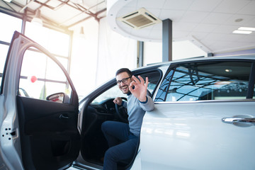 Car dealer or car buyer sitting in brand new vehicle showing okay hand gesture. Satisfied customer at vehicle dealership showroom.