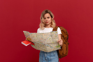 Thoughtful blond girl with curly hair in a white t-shirt looks at the map trying to find itinerary holding brown backpack