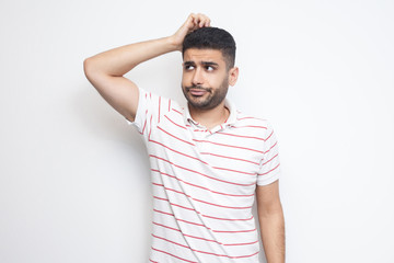 Portrait of query handsome bearded young man in striped t-shirt standing scratching his head, planing and thinking about something. indoor studio shot, isolated on white background.