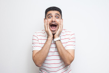 Portrait of surprised handsome bearded young man in striped t-shirt standing with hands on face and looking at camera with amazed face. indoor studio shot, isolated on white background.