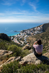 Naklejka premium Beautiful woman sitting on a cliff with the mediterranean sea and small coast town in the background