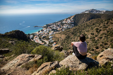 Naklejka na ściany i meble Beautiful woman sitting on a cliff with the mediterranean sea and small coast town in the background