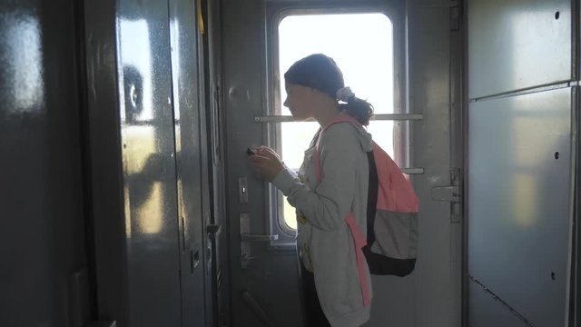 teenager girl traveler with backpack stands by the window of the train car with a smartphone. travel transportation railroad concept. the girl in the train at the window corresponds lifestyle the girl