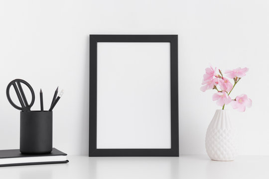Black frame mockup with pink oleander in a glass vase and workspace accessories on a white table.Portrait orientation.