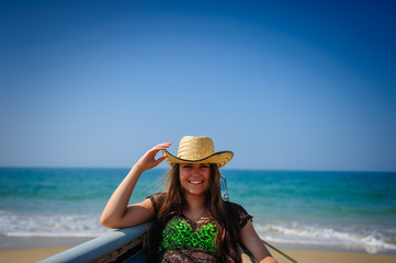 Portrait of young laughing girl with beautiful white teeth on a background of sandy beach, turquoise sea and bright blue sky. Pretty woman in straw hat, close-up.