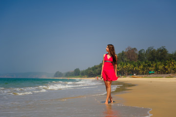 Young slender long-haired brunette in red dress walks barefoot on tropical beach along the surf against the sea and blue sky on a hot day. In the distance a green hill and people on the shore.