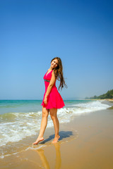Fototapeta na wymiar Young slender long-haired brunette in red dress standing barefoot on tropical beach with yellow sand against the sea and blue sky on a hot day. Vertical photo of beautiful girl walking along the surf.