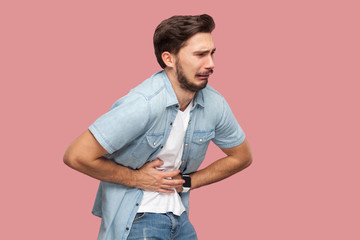 Stomach pain. Portrait of sick sad handsome bearded young man in blue casual style shirt standing...