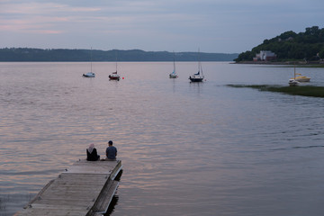 Fototapeta na wymiar Muslim couple sitting on a wooden jetty enjoying a summer blue hour evening on the St. Lawrence River shore in the Cap-Rouge bay area, Quebec City
