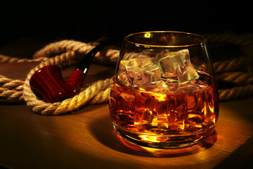 Glass with whiskey, ice cubes and smoking pipe on wooden background.