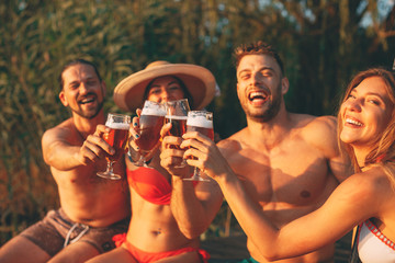 Happy group of young people toasting with beer on a dock by the river during the summer sunny day. Selective focus on beer