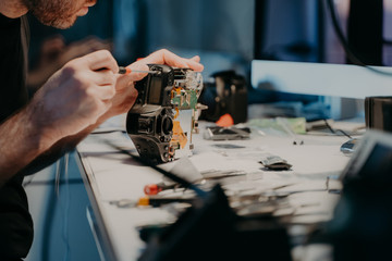 Woking process. Unknown man repairs professional digital camera, uses screwdriver, poses at...