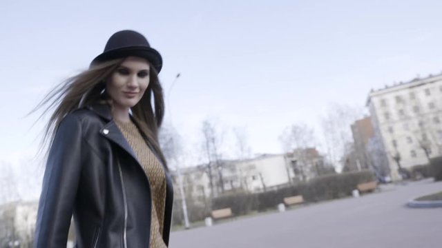 Woman walking along bare trees in the alley and people with children on the background. Action. Young model girl in a city park in autumn wearing black leather jacket and a hat.