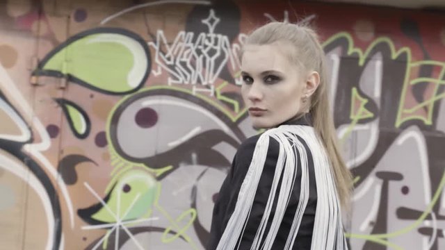 A portrait of a sexy brown haired girl with a ponytail wearing stylish black jacket with white long fringe standing against graffiti wall background. Action. Model woman in urban environment.