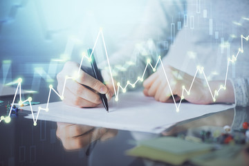 Hands writing information about stock market in notepad. Forex chart holograms in front. Concept of research. Multi exposure