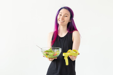 Slim young asian girl with colored hair is holds a measuring tape and holding a light vegetable salad in her hands posing on a white background. Healthy eating concept. Copyspace.