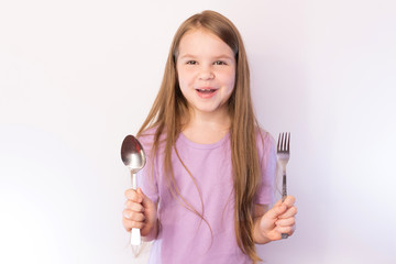 Little cute girl holding a spoon and fork with a satisfied expression on her face, on a light background