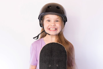 Cute little girl in protective helmet with a funny expression holding a skateboard against a bright background