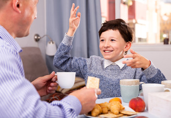 Son talking with father at table with tea during breakfast indoors