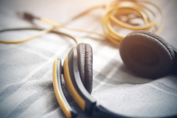 Yellow-black headphones with a microphone, the wire which is tangled in a ball, lie on a gray blanket in a white strip and illuminated by sunlight.