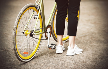 Fototapeta premium A girl in black pants and white sneakers stands near her beautiful bike is yellow-green in the Park in the summer.