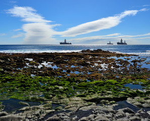 Rocky beach at low tide in foreground, blue sky with peculiar cloud and ships in the bay 