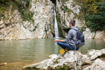 Young male travaler in casual clothes with backpack sits in picturesque place on stone overlooking...