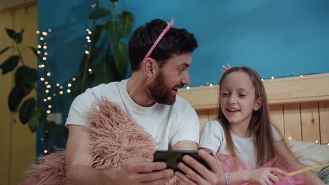 happy father and daughter dressed magic fairies playing together watching videos on smartphone. cute girl waving with magic wand sitting on bed with daddy at home. birthday party.