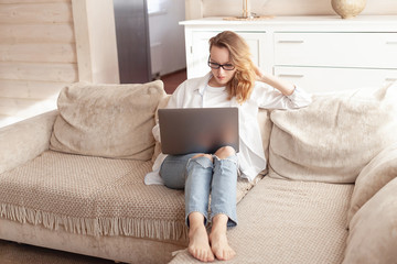 Serious young pretty businesswoman working remotely using her laptop sitting on a sofa in the living room of her cozy country house