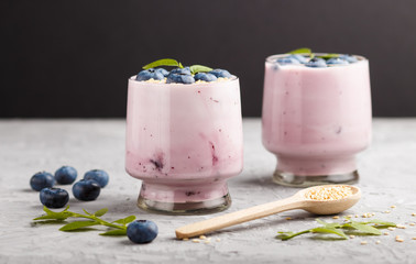 Yoghurt with blueberry and sesame in a glass and wooden spoon on gray and black background. side view.