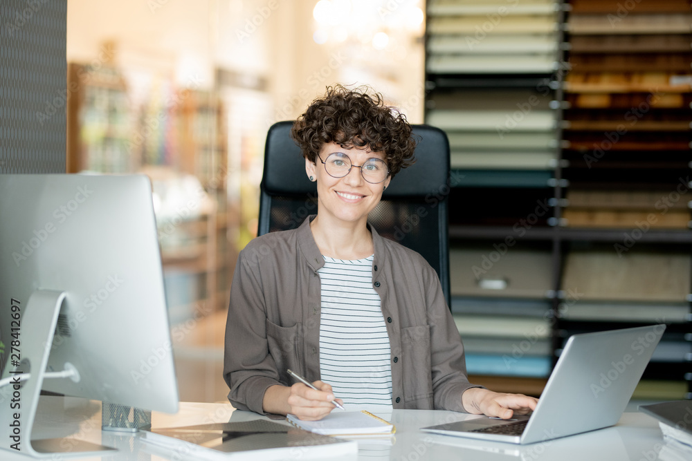 Poster young attractive curly designer making notes in front of laptop