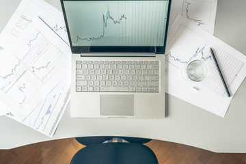 Top view. Young business woman sitting at table with cup of coffee and working on the laptop with graphics and charts printed on the paper. 
