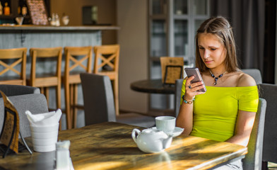 portrait of young teenager brunette girl with long hair sitting indoor in urban cafe and use her smartphone