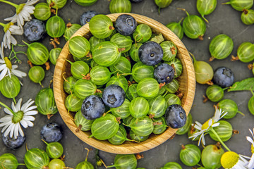 Top view on the berries of gooseberry and blueberry in a wooden bowl. In the center of the dark table is a cup with green and blue berries, around which these berries are scattered. 