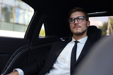 Thoughtful confident businessman in full suit with eyewear looking away while sitting in the car.