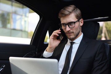 Handsome confident businessman in suit talking on smart phone and working using laptop while sitting in the car.