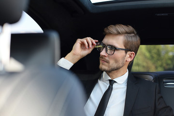 Thoughtful confident businessman keeping hand on glasses while sitting in the luxe car.
