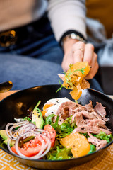 Woman eating salad with avocado, tomatoes, poached egg, beef, orange, onion and arugula. Salad in a black plate on a yellow set in a restaurant. Close-up. space. photo series
