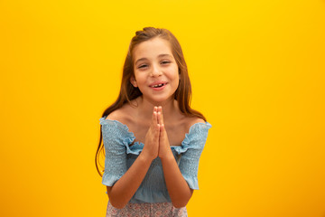 Nice little girl, holds hands in praying gesture, stands over isolated yellow background with copy...