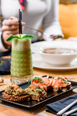 Whole grain bread with avocado paste mussels and shrimp or salmon and cheese on a black plate and a glass of green smoothie on a table in the restaurant. in the background a girl eats soup. close up