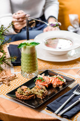 Whole grain bread with avocado paste mussels and shrimp or salmon and cheese on a black plate and a glass of green smoothie on a table in the restaurant. in the background a girl eats soup. close up