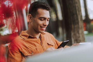 Joyful young man in orange shirt using cellphone outdoors