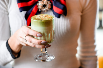 Woman in a light sweater holds a beautiful faceted glass of green matcha latte, sprinkled with nut chips. close-up. Concept - healthy food