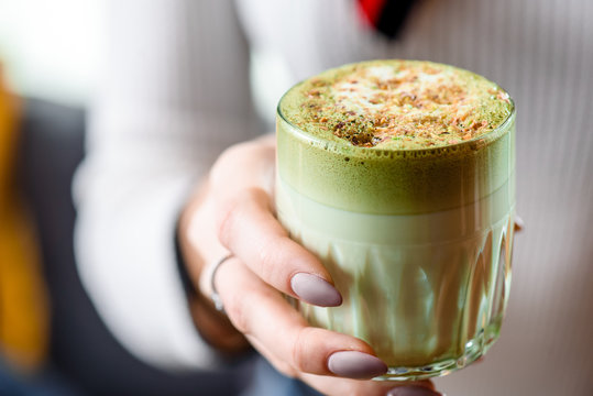 Woman in a light sweater holds a beautiful faceted glass of green matcha latte, sprinkled with nut chips. close-up. Concept - healthy food