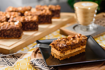 Chocolate brownie with nuts on a black plate on a yellow set on a wooden table in a restaurant. Cappuccino on the background. Close up. Space