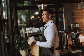 Cheerful barman wiping white cup with cleaning cloth