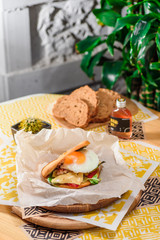 Burger with fried egg, chicken, cheese, tomatoes and other vegetables on a craft paper on yellow background in a restaurant. Toasted black bread on the background. Series of photos for the menu.