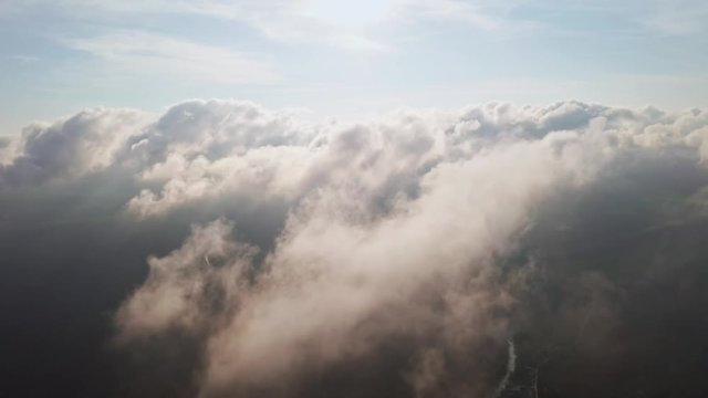 Through moving white clouds on coastline and sea surface scenic aerial view. The drone flies forward high in the blue sky through the fluffy clouds on panorama of sea shore. The sun is hidden. Halo