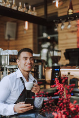 Handsome barman with cup of coffee using cellphone in cafe
